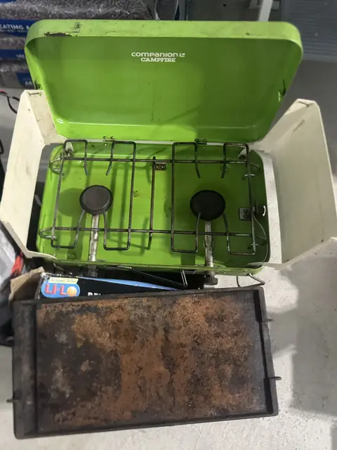 A green two-burner camping stove with its lid open, and a cast iron hotplate in front.