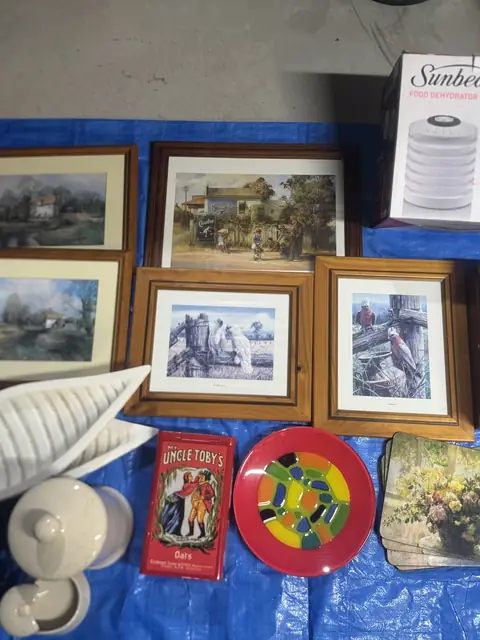 Framed country and bird prints, an Uncle Toby&rsquo;s oats tin, a bright glass platter and a stack of floral placemats on a tarp.