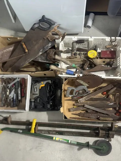 Boxes of old hand tools: rusty saws, files, spanners, a brace and bit, a circular saw blade and an electric drill, with two long sash clamps in front.
